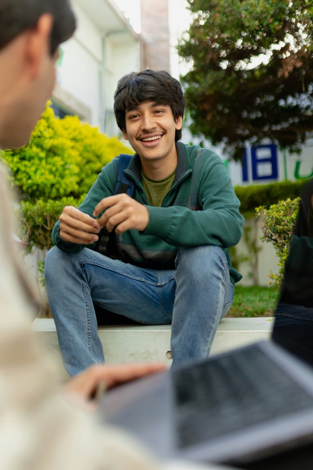A smiling teen sits on outdoor steps talking with someone holding a laptop