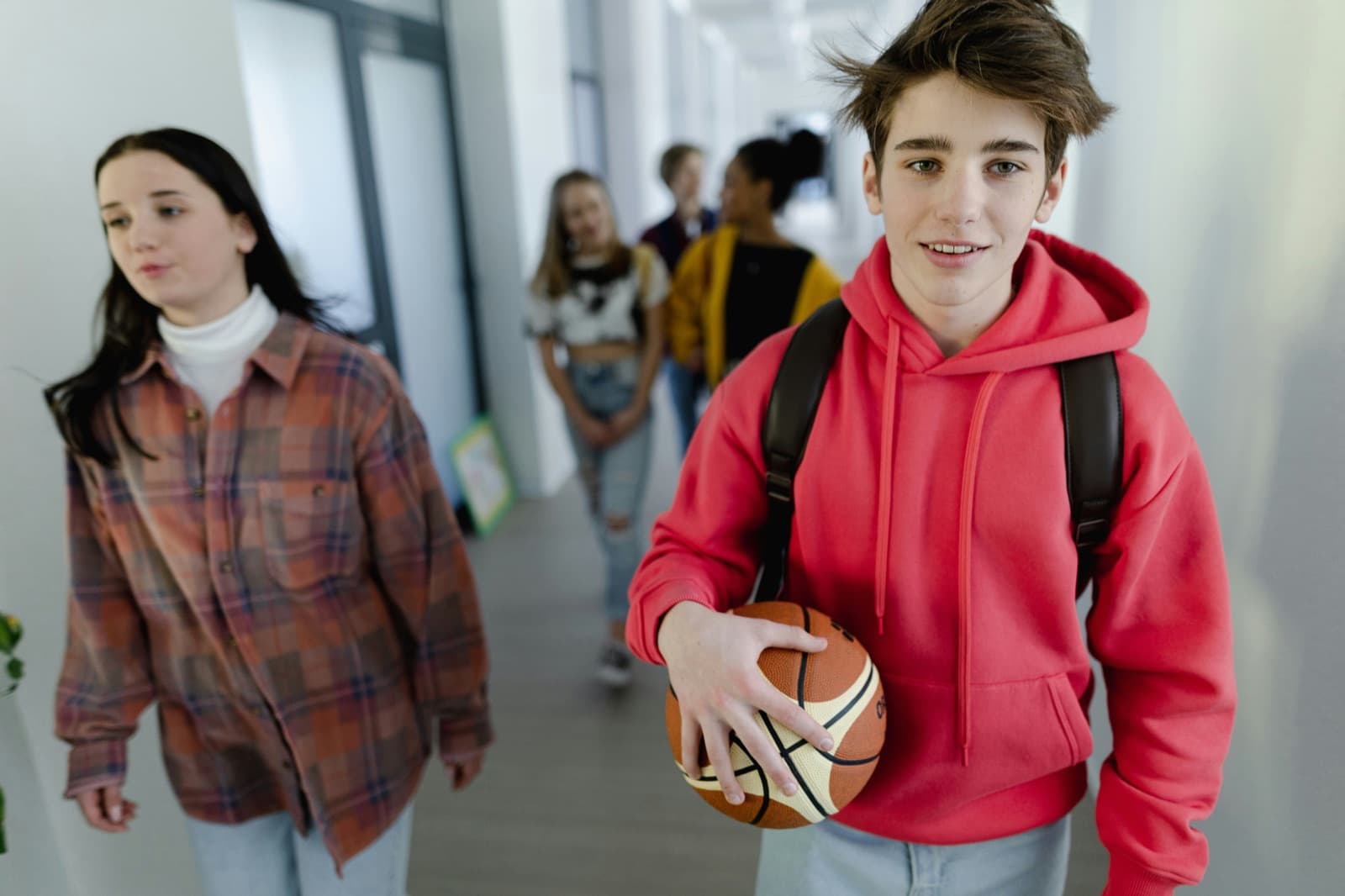 A teen carrying a basketball smiles while walking down a school hallway with classmates