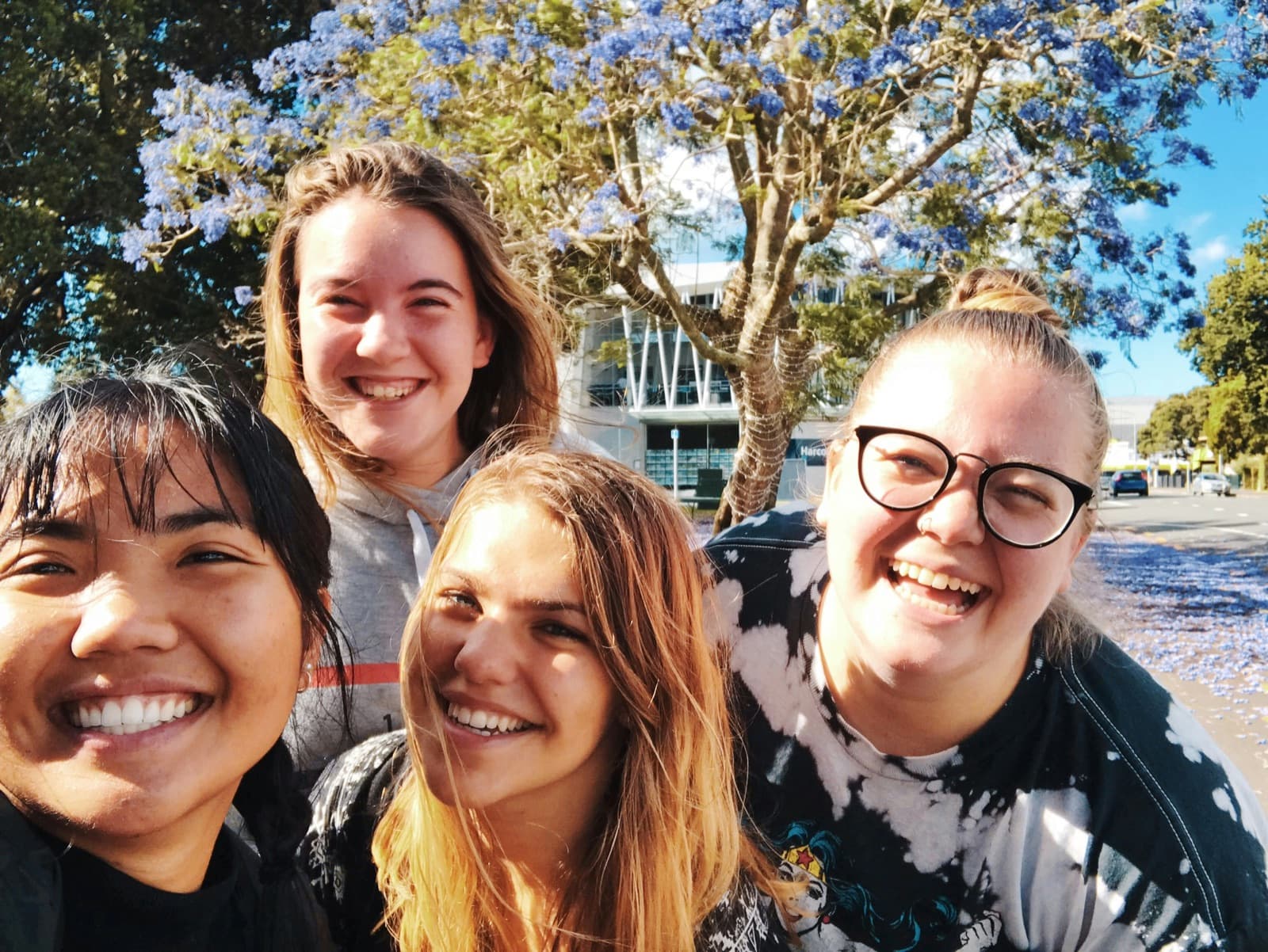 Four smiling students in a sunny selfie under a jacaranda tree on campus