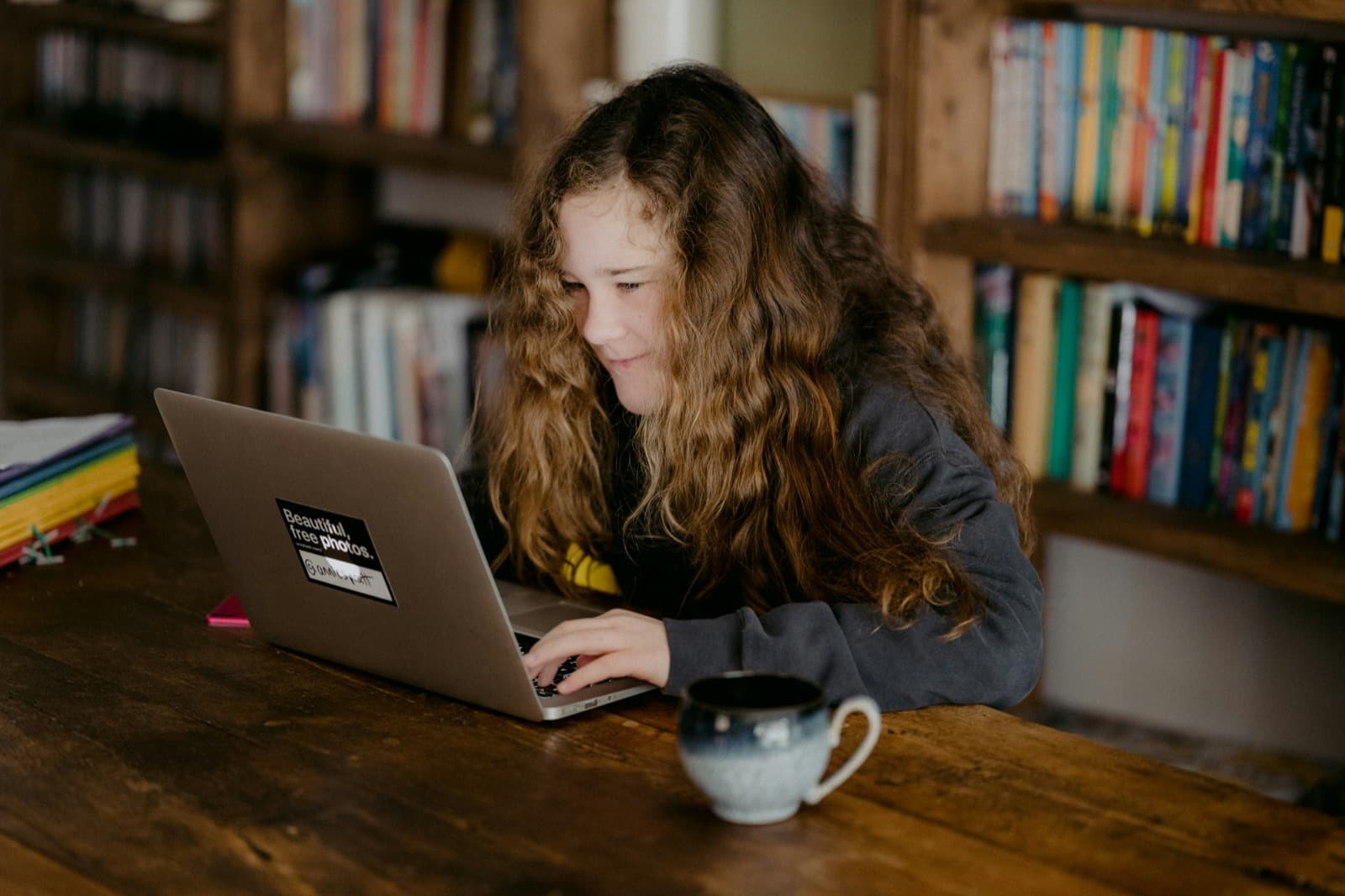 A teen with long curly hair smiles while working on a laptop in front of bookshelves