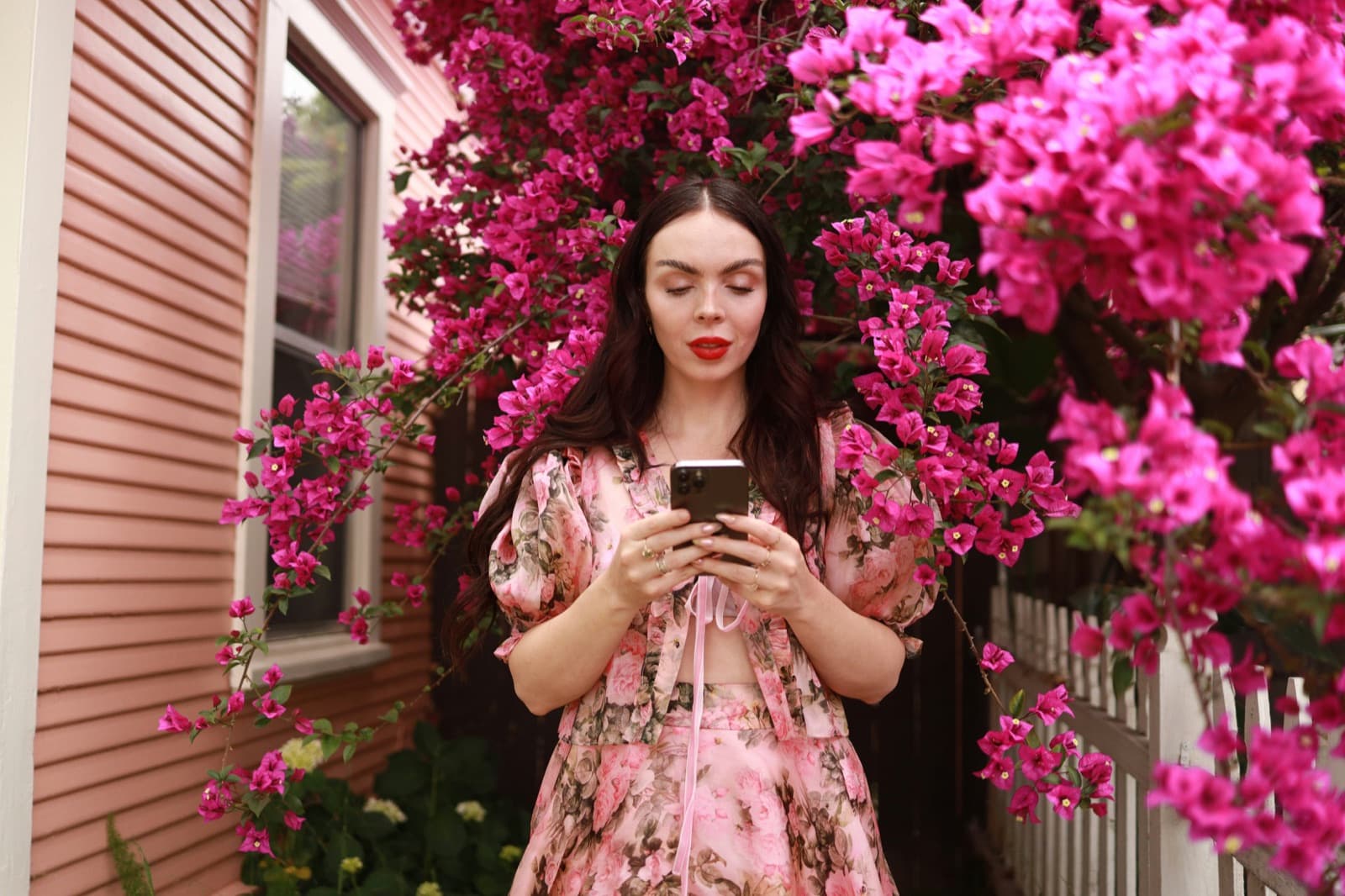 A young person checks their phone beside a house covered in bright pink flowers