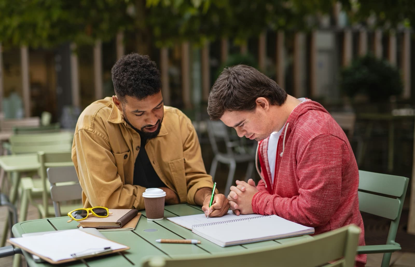 Two teens studying together at an outdoor café table with notebooks and coffee