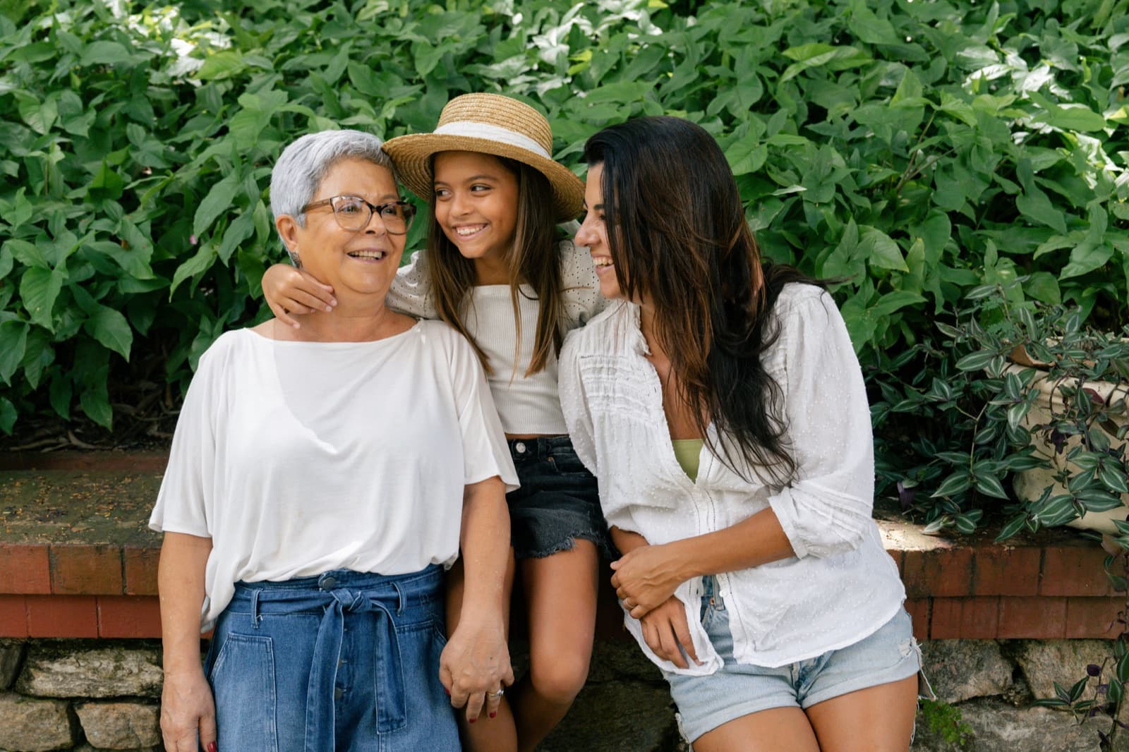 Three generations of women smiling together in front of leafy greenery