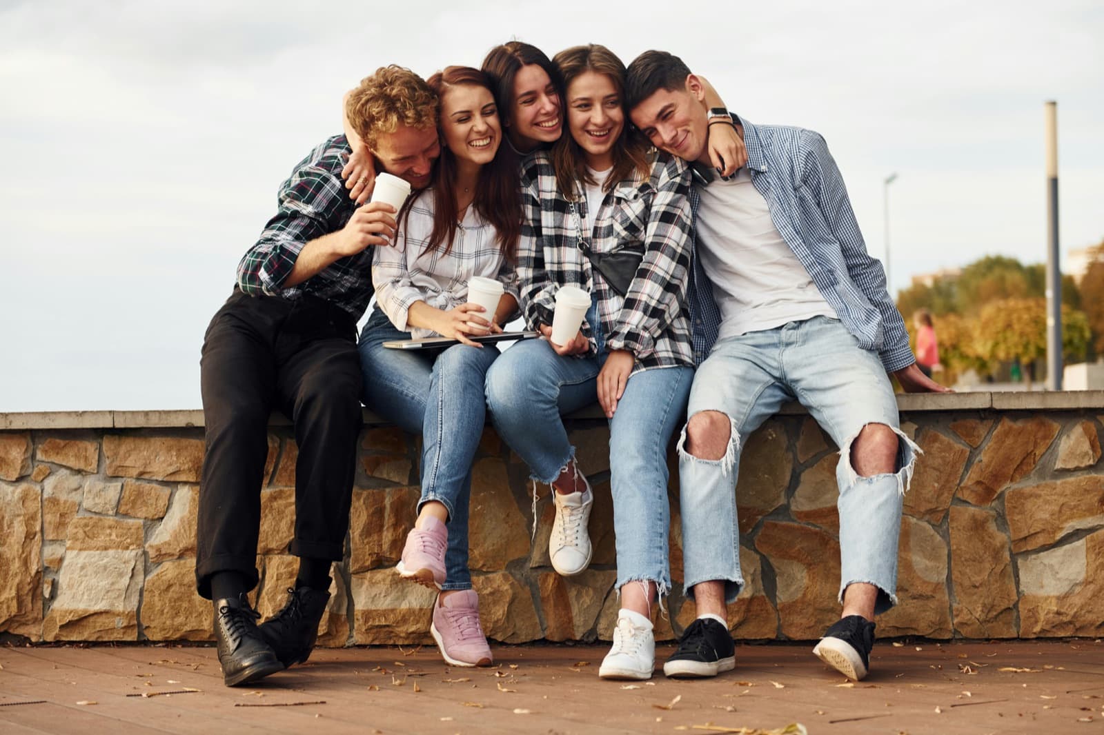 Five young adults sitting close together on a low wall, laughing